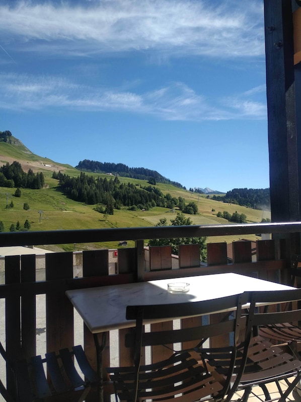 A balcony with a table and chairs, overlooking a green landscape with hills and trees.