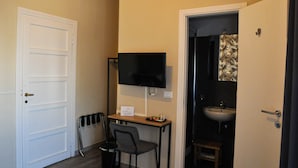 Ein kleines Zimmer mit weißer Tür, einem Schreibtisch mit Stuhl, einem Flachbildfernseher, einem Waschbecken und einem Spiegel.