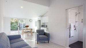 Ein modernes Wohnzimmer mit grauem Sofa, einem grauen Sessel und einem Essbereich mit Tisch und Stühlen. Es gibt eine Küchenzeile mit weißen Schränken und einer Mikrowelle. Eine Glastür öffnet sich zum Garten.