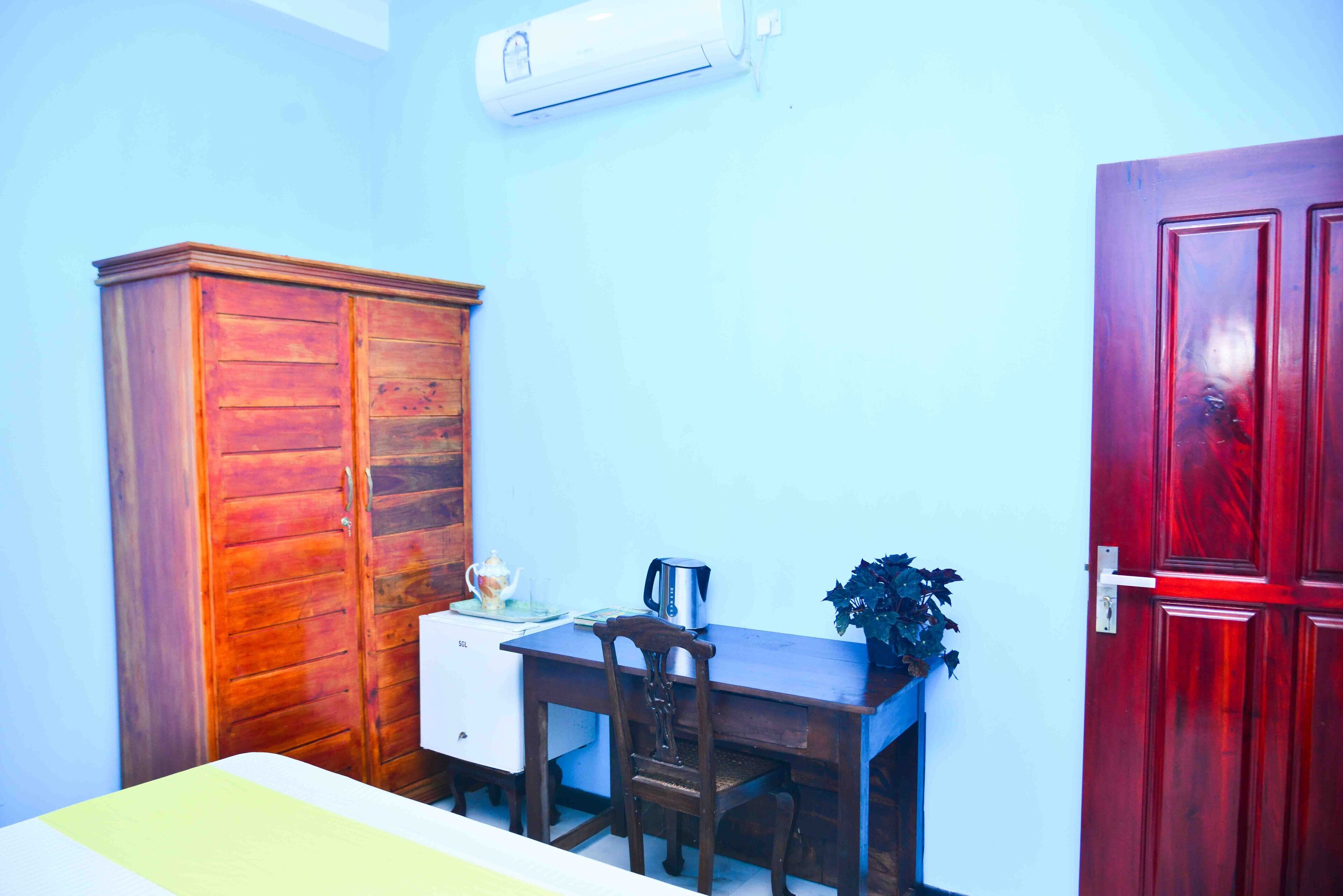Een kamer met een houten garderobe, een bureau met stoel, een kleine tafel met een plant, en een rode deur.
