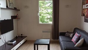 Un salon équipé d’un téléviseur à écran plat, d’un canapé noir avec des coussins, d’une petite table noire et d’une fenêtre avec des rideaux.
