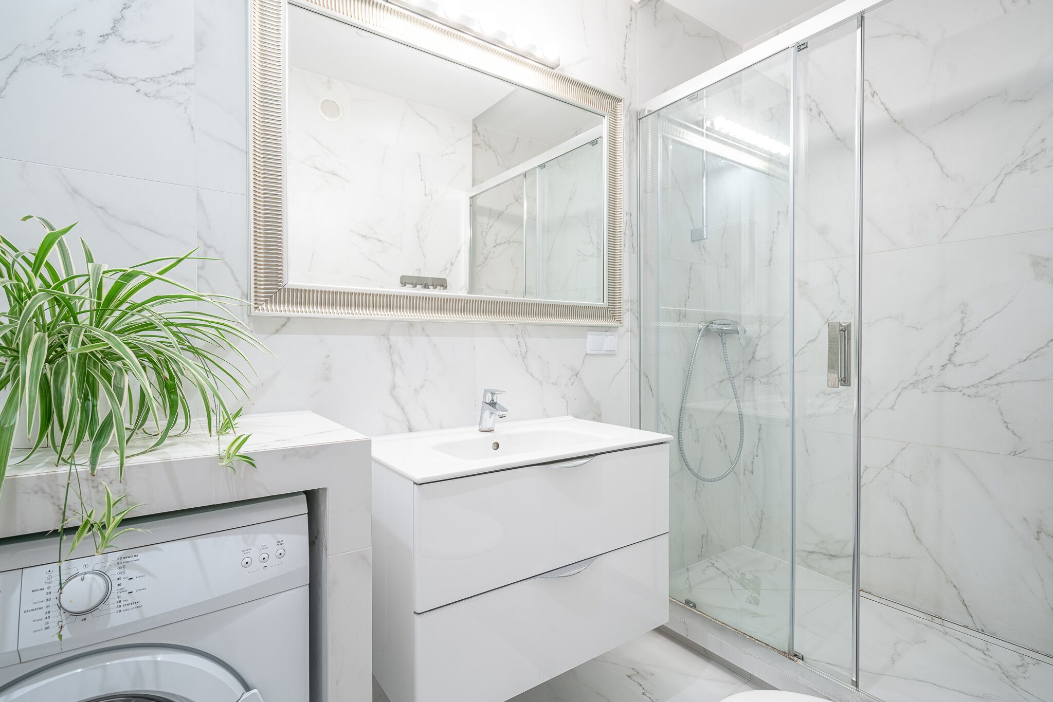 A modern bathroom with a marble sink, a large mirror, a glass shower enclosure, and a washing machine.