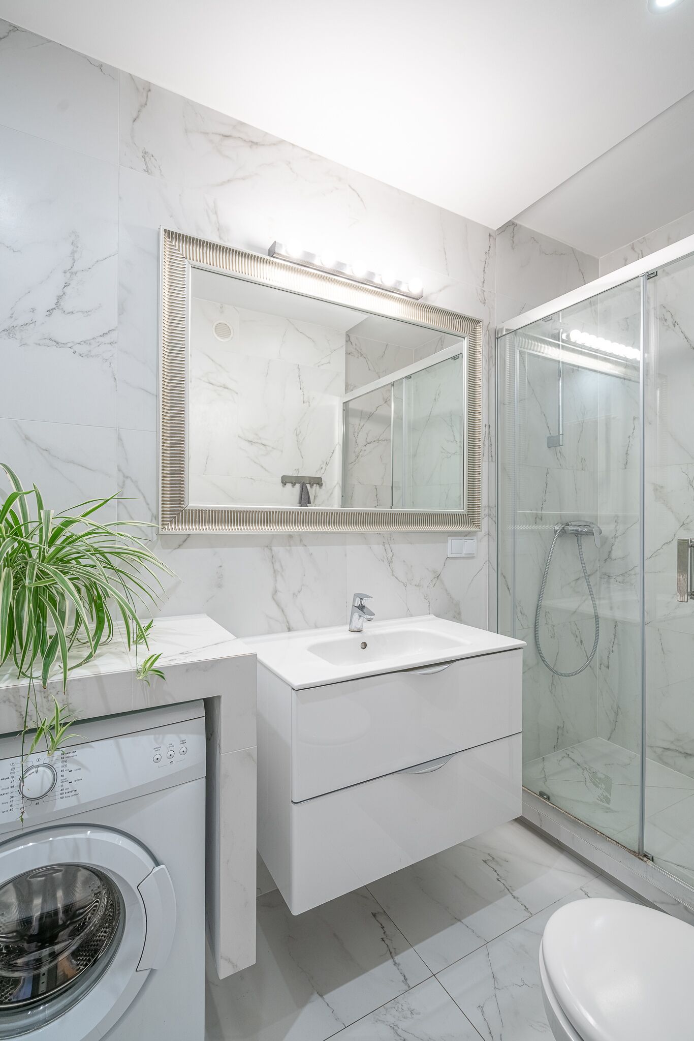 A modern bathroom with a marble backsplash, a large mirror, a shower enclosure, and a built-in washing machine.