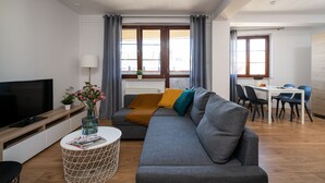 A modern living room with a grey sofa, a flat-screen TV, a coffee table, and a dining area with a white table and blue chairs. - LeoApart (Wroclaw)