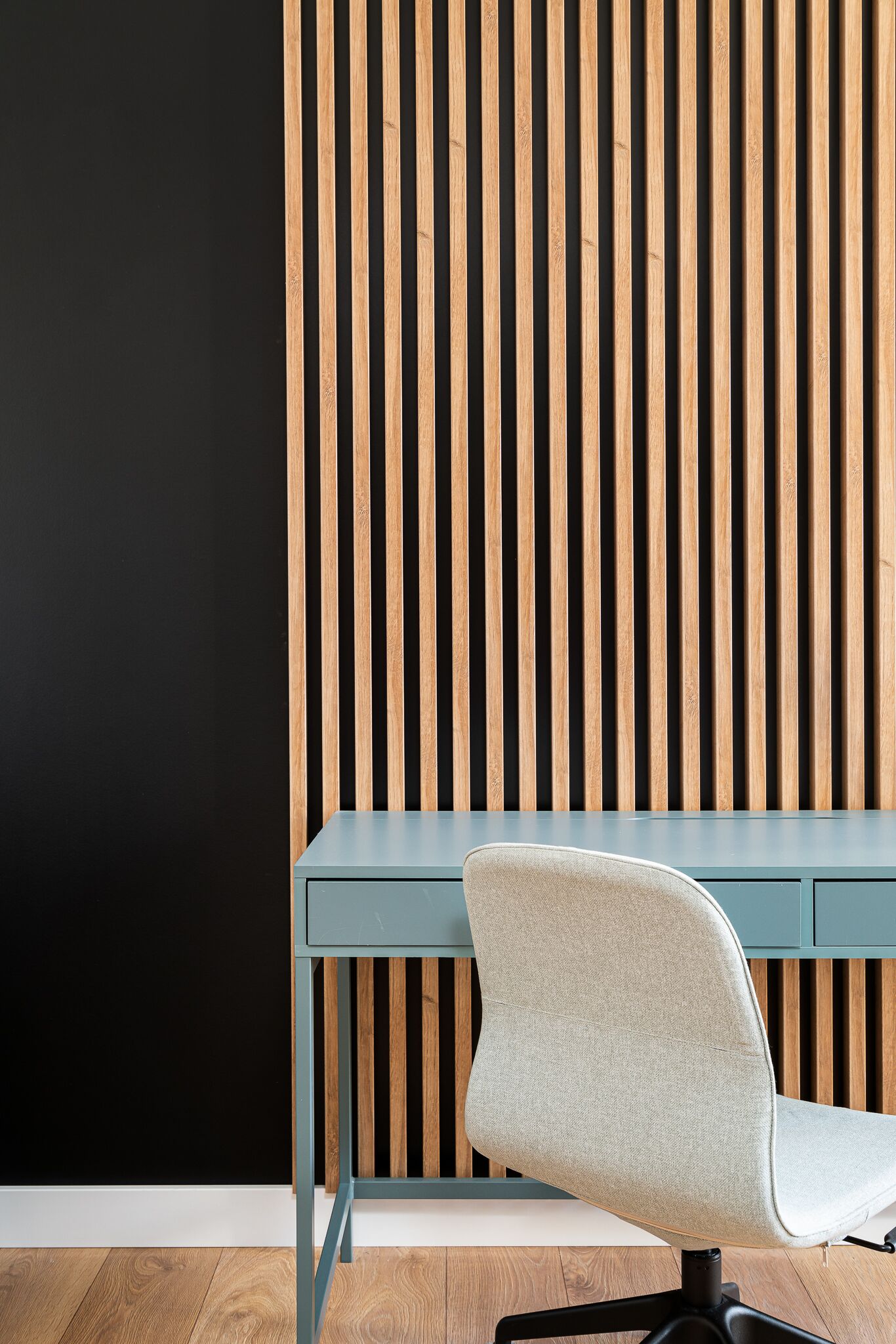 Una silla de oficina moderna con asiento y respaldo de tela gris claro, ubicada frente a un escritorio azul oscuro sobre un fondo de listones de madera verticales.