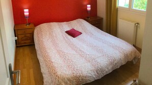 A bedroom with a red wall, a bed with a white and red patterned bedspread, two bedside lamps, and a window with a view of greenery.