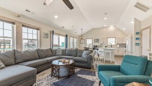 A spacious living room with a large sectional sofa, a teal armchair, and a coffee table. The room has large windows, a ceiling fan, and a kitchen area visible in the background. - Renovated - Seaside 5BR Caribbean Escape near the Rodanthe Pier (Rodanthe)