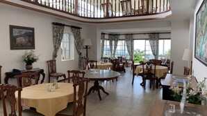 Un vaste espace de salle à manger avec plusieurs tables et chaises rondes, un balcon doté de rampes en bois et de grandes fenêtres offrant une vue sur la verdure.
