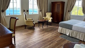 Une pièce avec un plancher en bois, un lit, deux chaises, une armoire en bois et deux fenêtres avec des rideaux.