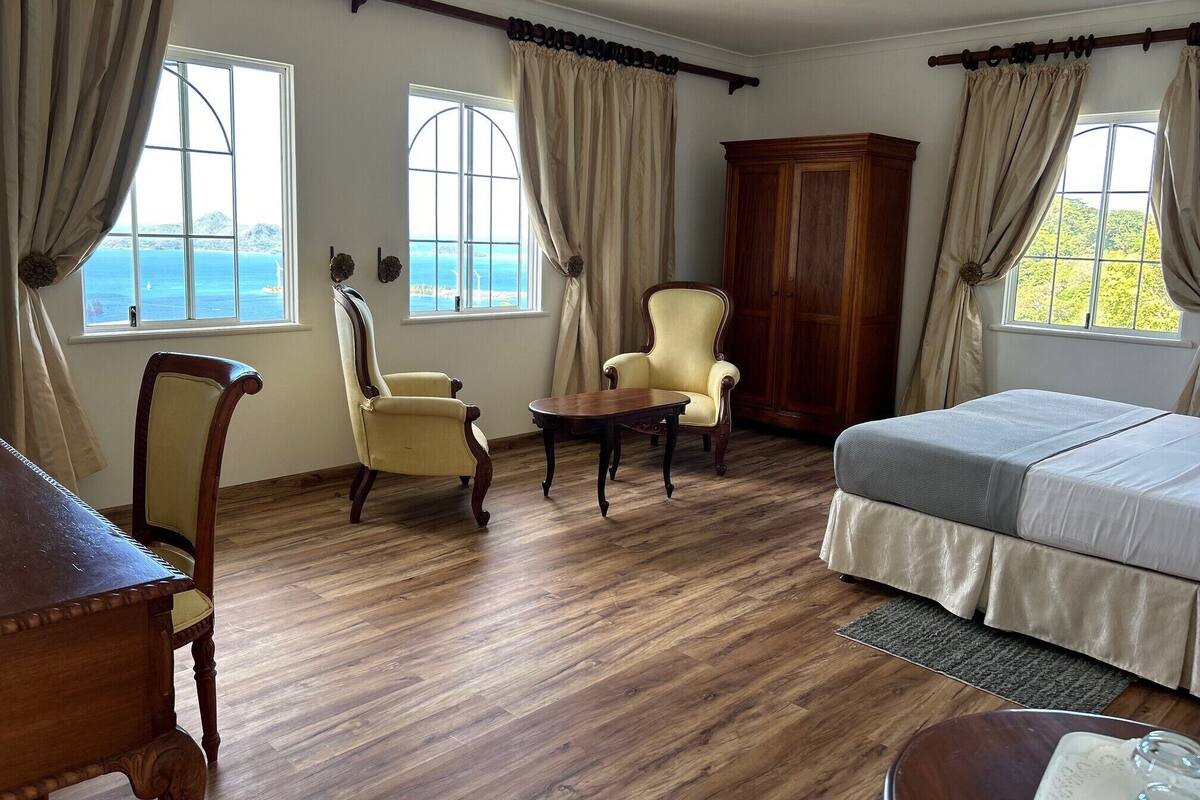 Ein Zimmer mit Holzboden, einem Bett, zwei Stühlen, einem Holzschrank und zwei mit Vorhängen versehenen Fenstern.
