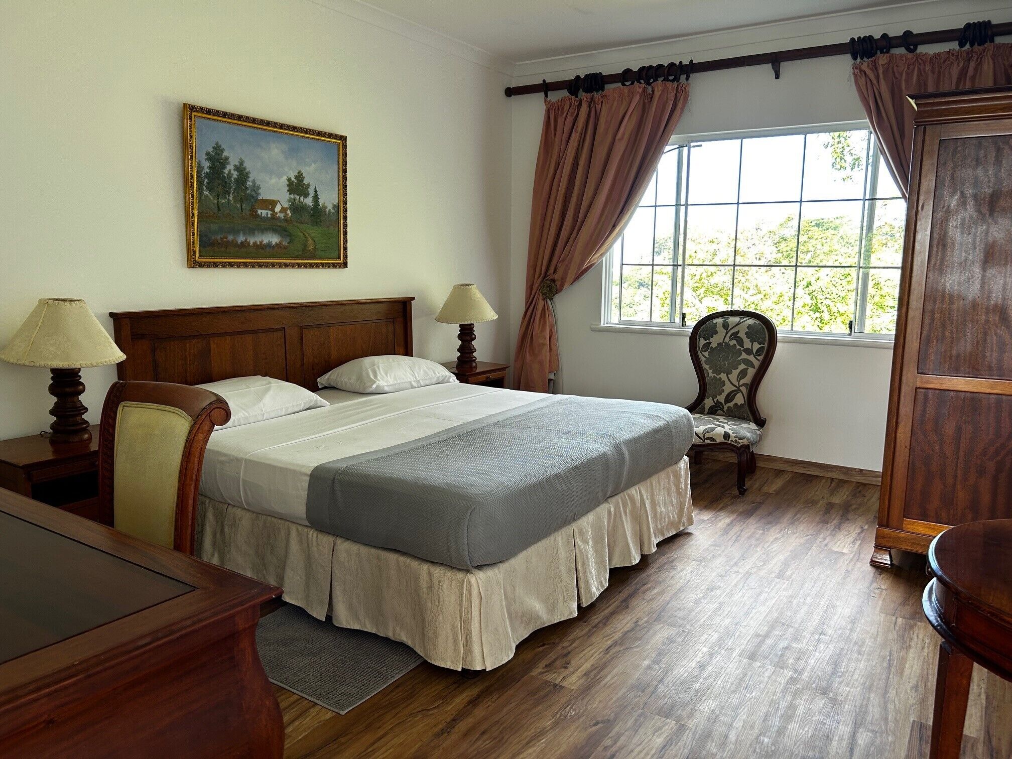 A neatly arranged bedroom with a bed, two bedside tables, a chair, a window with curtains, and a painting on the wall.