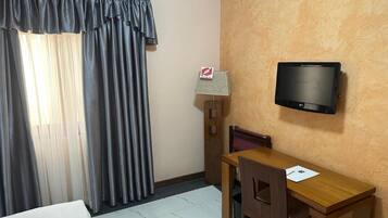 A hotel room with a bed, a desk, a chair, a television mounted on the wall, and a window with curtains.