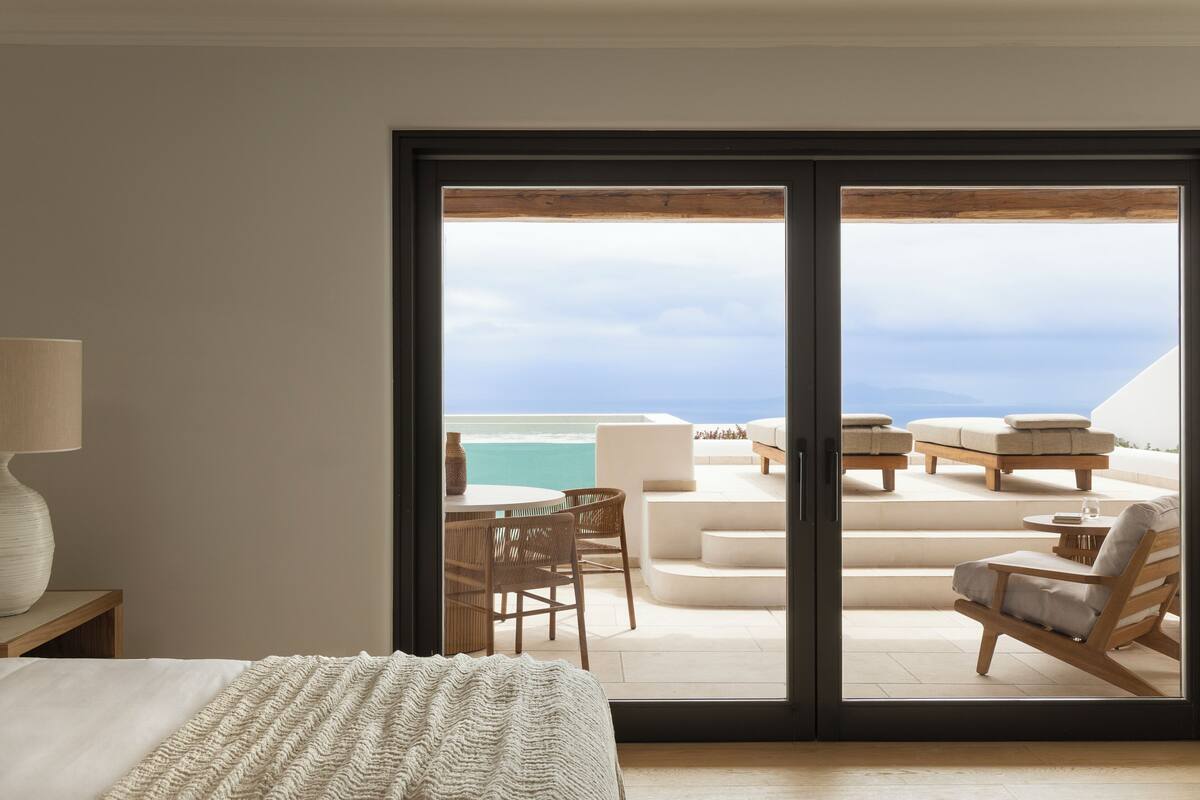 A bedroom with a large sliding glass door leading to a balcony with outdoor seating and a view of the ocean.