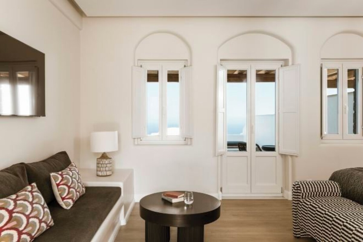 A living room with a sofa, a round coffee table, and a patterned armchair. There are three arched windows with white frames and a view of the sea.