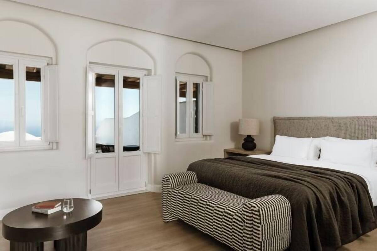 A bedroom with a bed, a bench, a round table, and two arched windows.