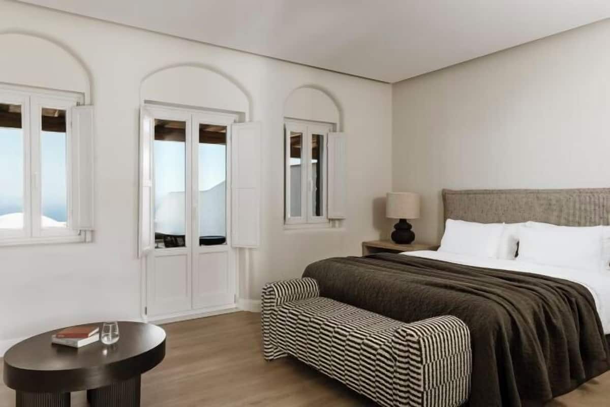 A bedroom with a bed, a bench, a round table, and two arched windows.