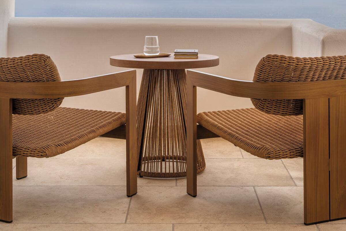 A wooden table with two wicker chairs, a glass, and a book on a balcony with a view of the sea.