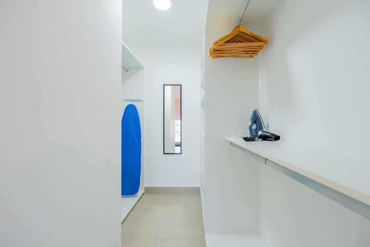 Un pasillo estrecho con una toalla azul colgada a la izquierda, un espejo en la pared y una estantería con un secador de pelo a la derecha.