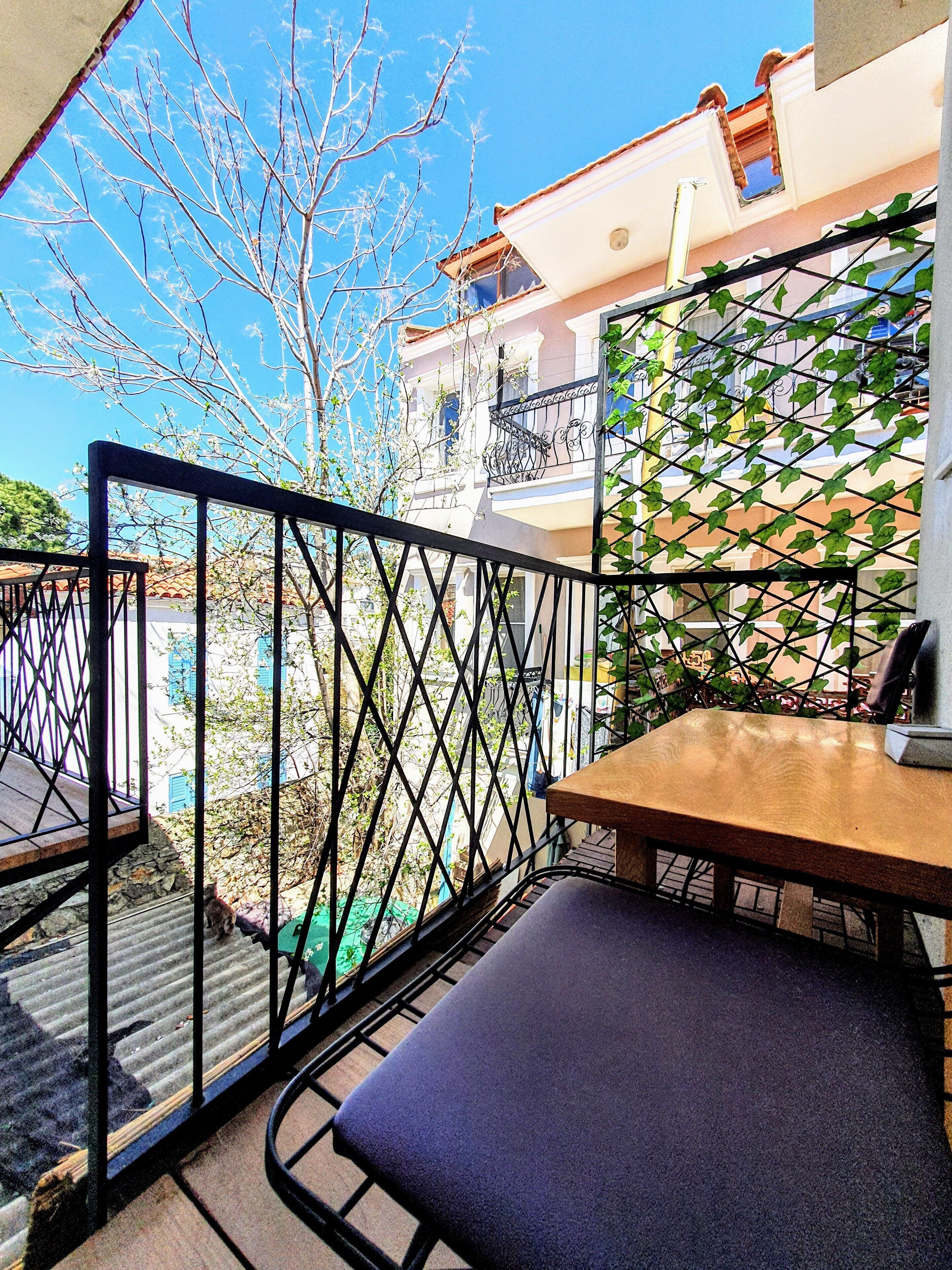 Un balcón con una mesa de madera y un banco acolchado, con vistas a una zona residencial con casas y un árbol.