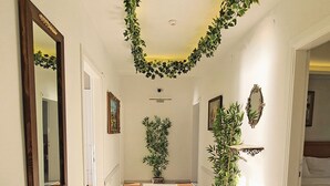 Un couloir aux murs blancs, doté d’un plancher en bois et d’un plafond orné de motifs végétaux verts. Des portes se trouvent de part et d’autre, un miroir est accroché à gauche et un lit avec une tête de lit se trouve à droite.