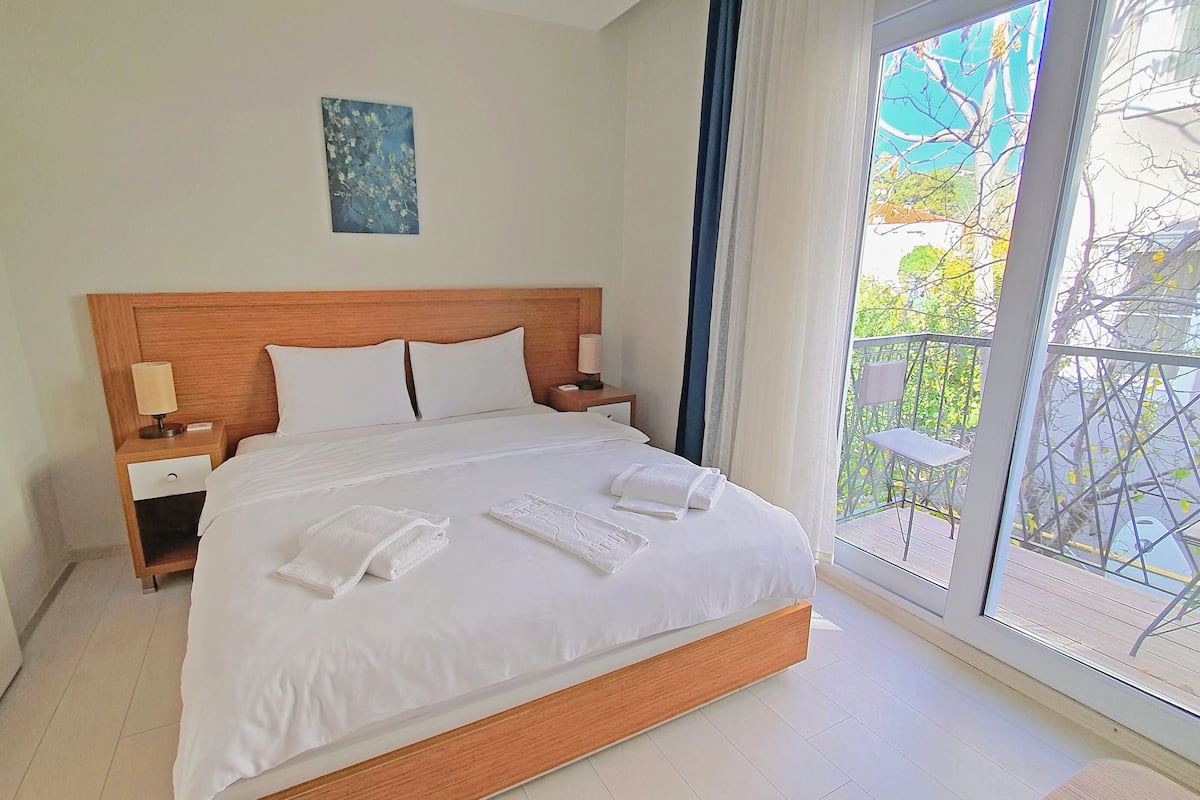 A hotel room with a large bed, a wooden headboard, two bedside tables with lamps, a painting on the wall, and a balcony with a view of trees and a chair.