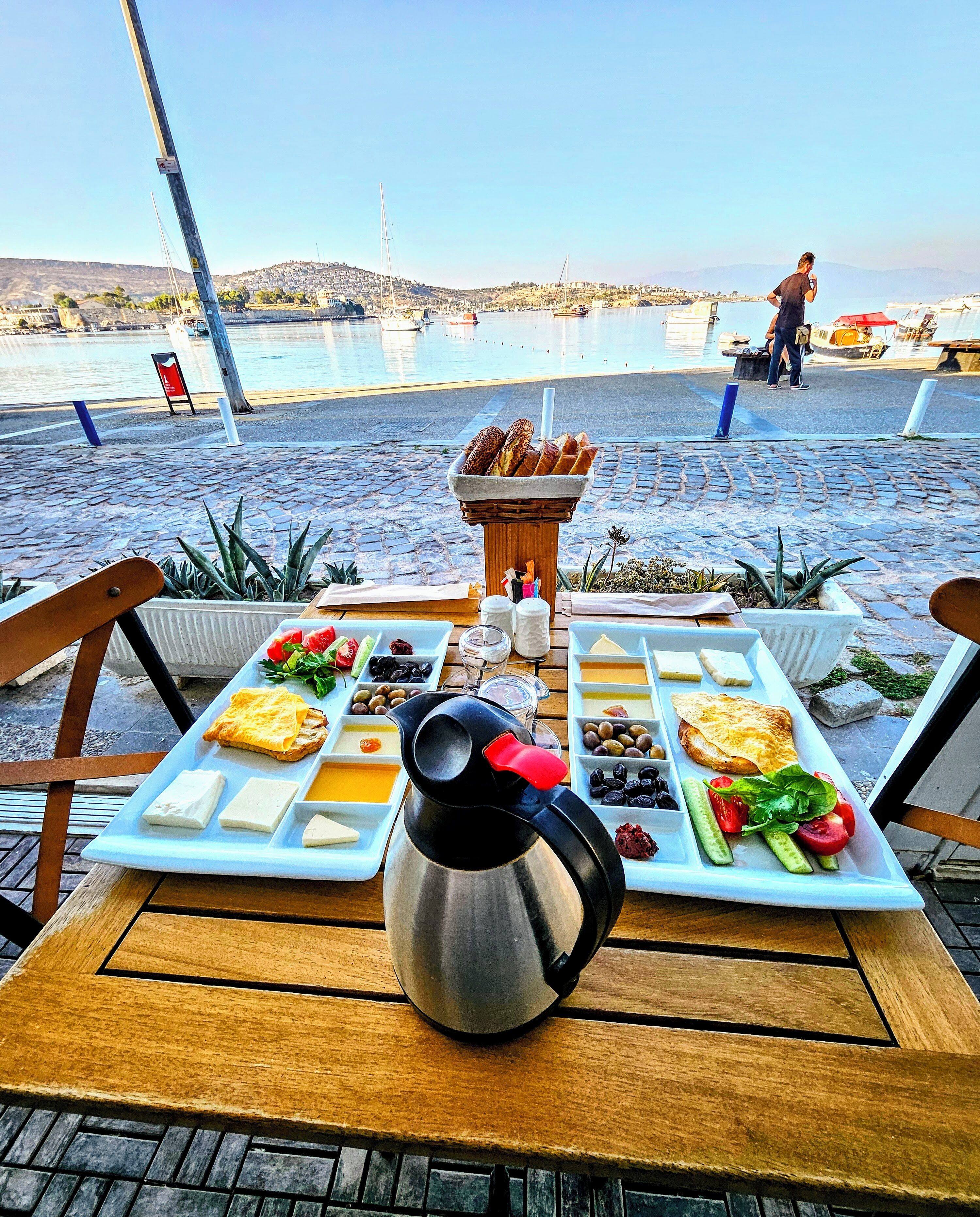 Ein gedeckter Tisch mit Frühstücksgebäck, Käse und Eiern, mit Blick auf Boote und Hügel im Hintergrund.