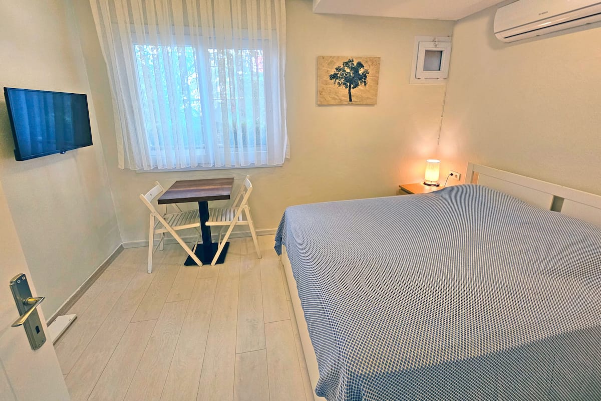 A bedroom with a bed, a desk, a chair, a window with curtains, a wall-mounted air conditioner, and a painting.