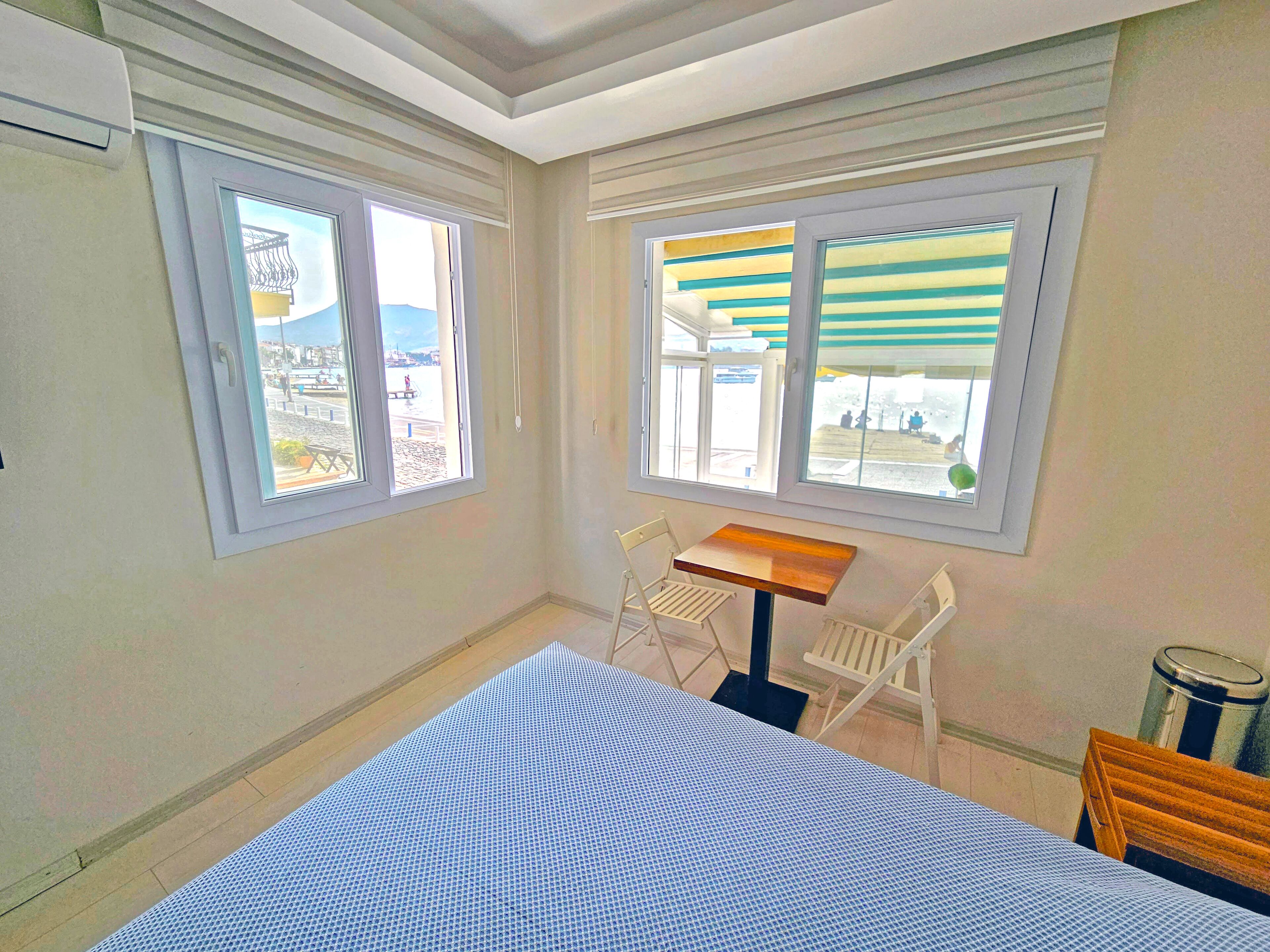 Ein kleines Zimmer mit einem Bett, einem Tisch und zwei Stühlen. Fenster bieten Ausblicke nach draußen.