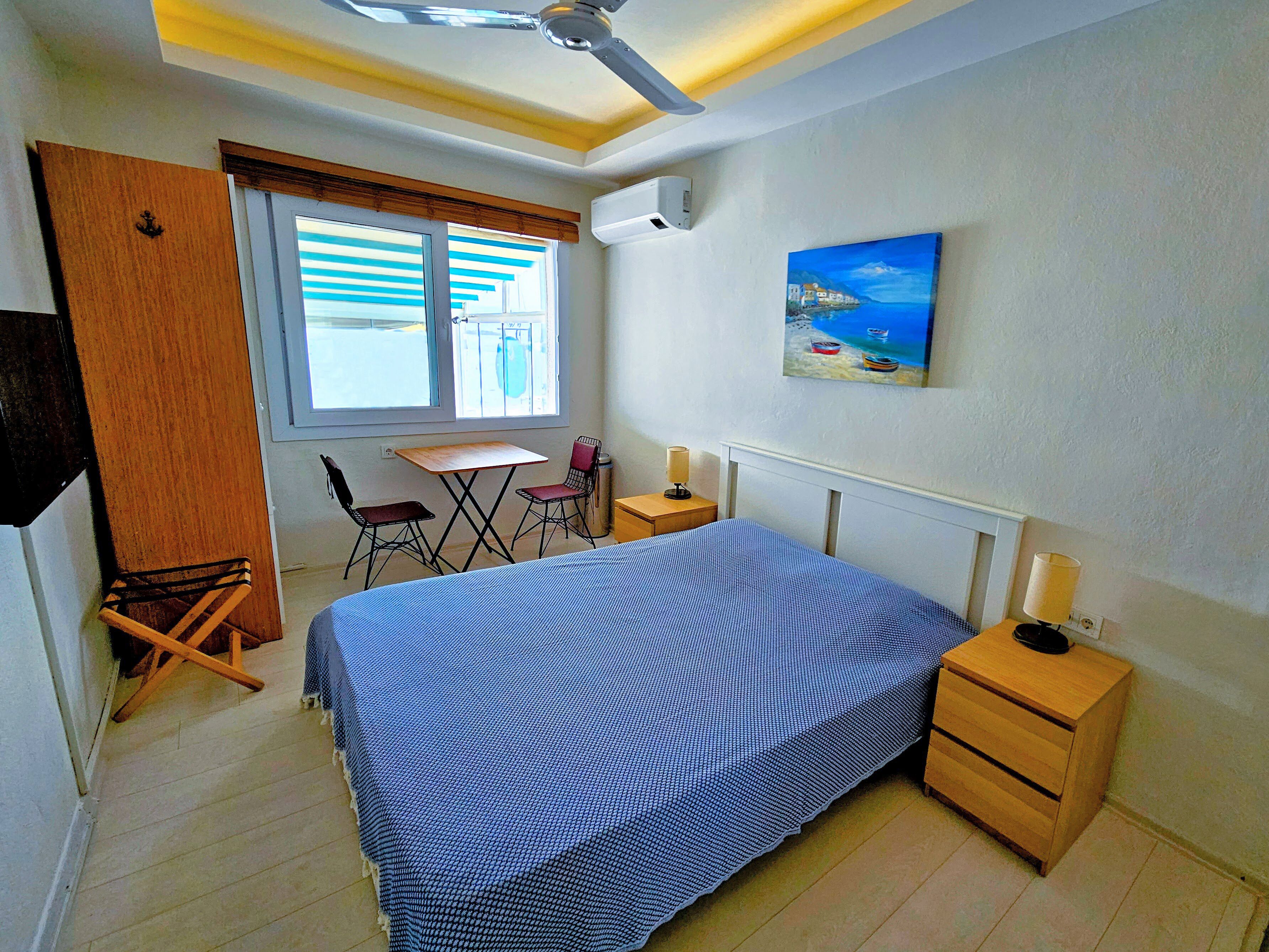 A bedroom with a bed, a desk, two chairs, a wooden wardrobe, a ceiling fan, a painting, and a nightstand with a lamp.