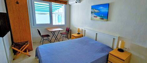 A bedroom with a bed, a desk, two chairs, a wooden wardrobe, a ceiling fan, a painting, and a nightstand with a lamp.