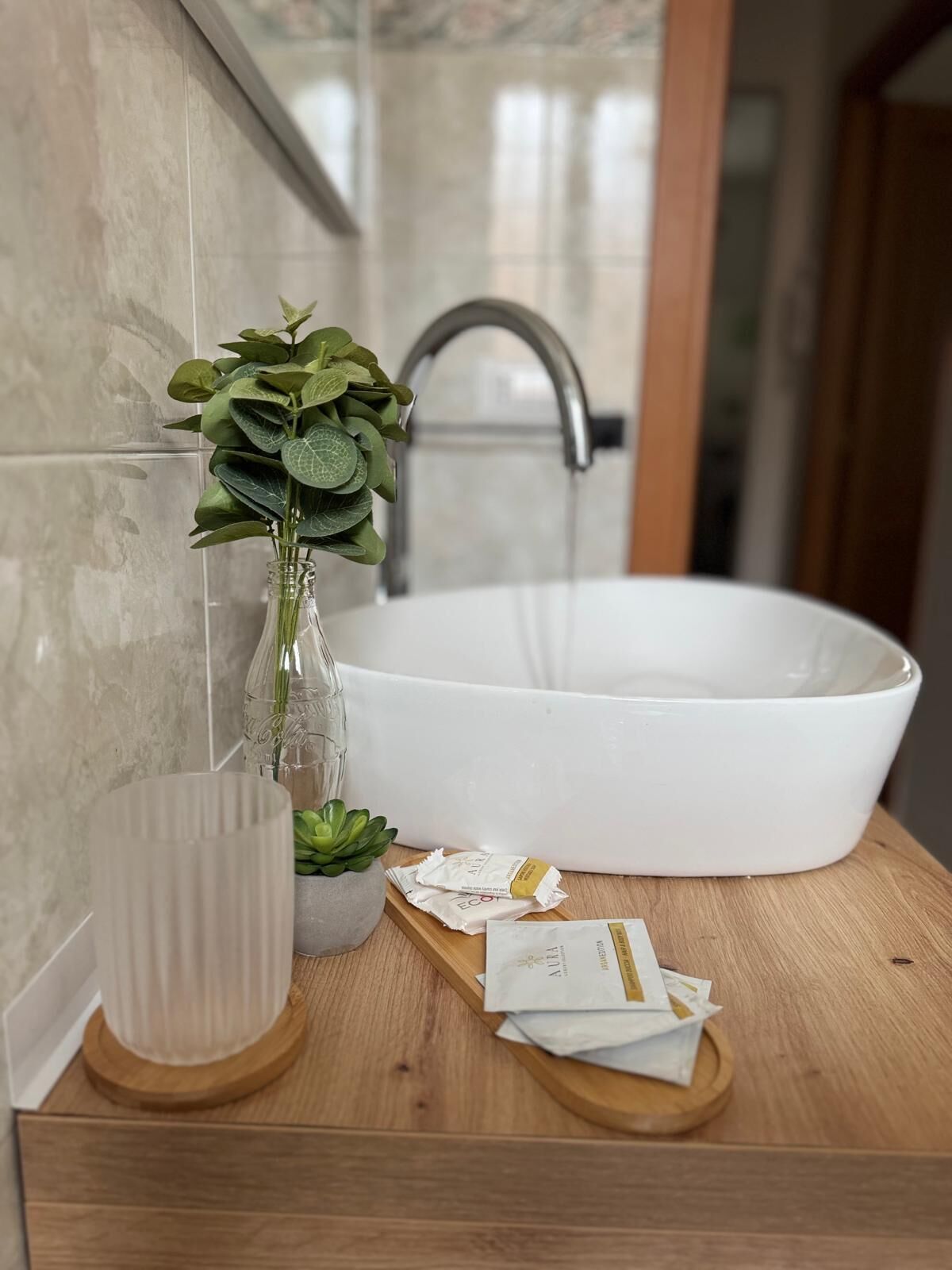 A bathroom with a white sink, a glass vase with green leaves, a small potted plant, and a wooden tray with toiletries.