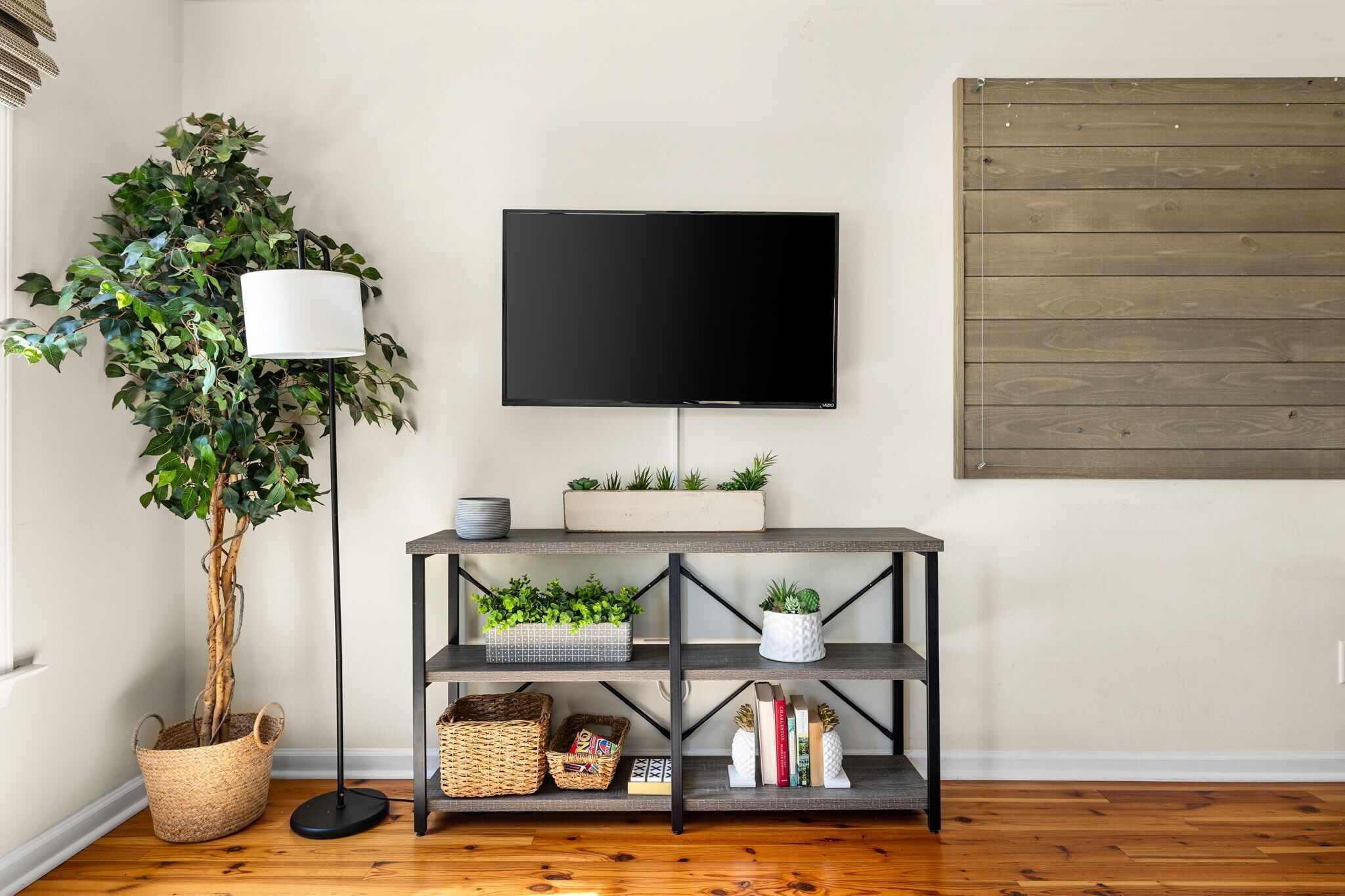 A living room with a TV mounted on the wall, a wooden floor, a potted plant, a floor lamp, and a console table with decorative items.
