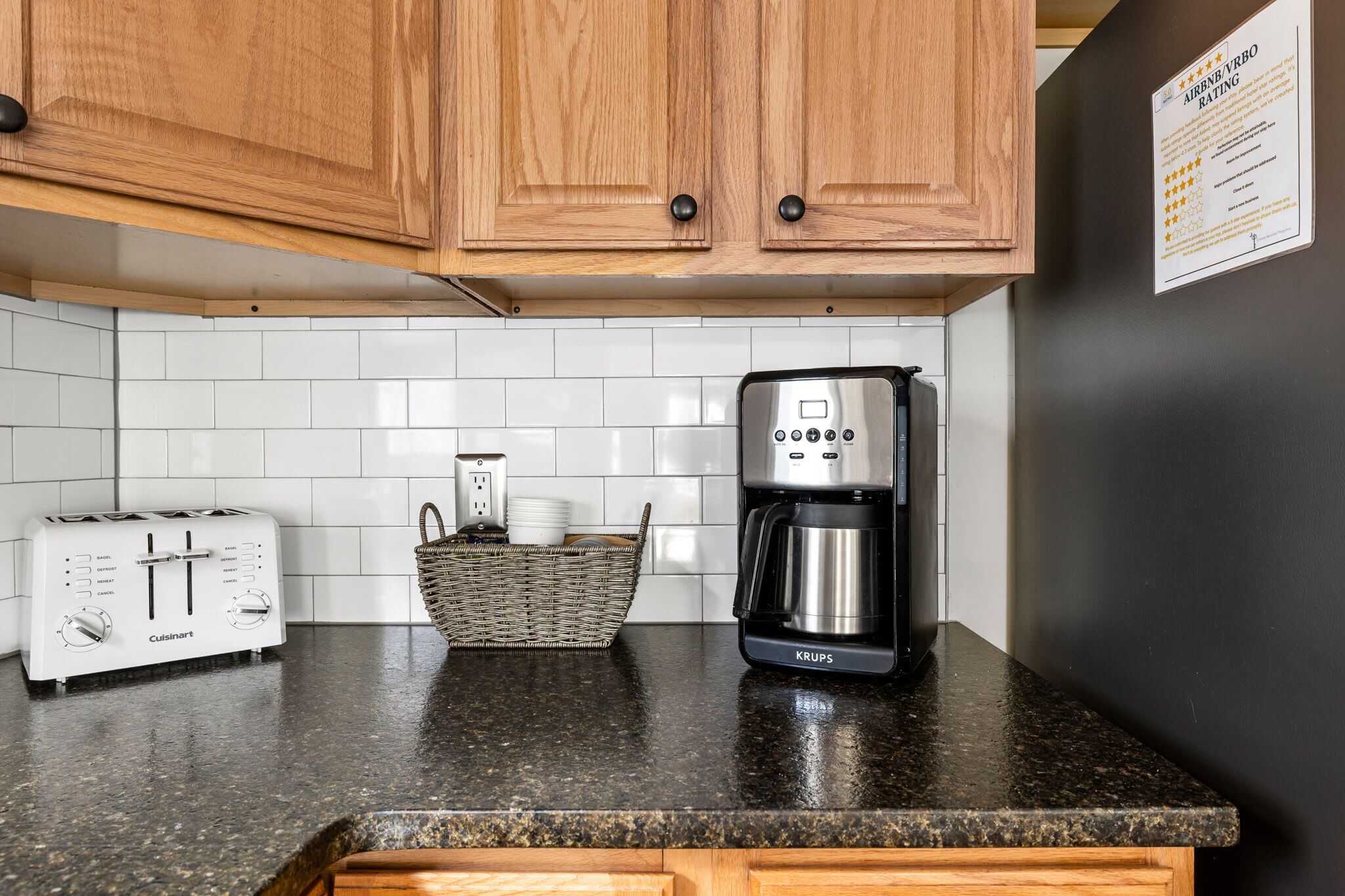 Una cocina con armarios de madera, salpicadero de azulejos blancos, encimera negra, un tostador Cuisinart, una cafetera Krups y una cesta de mimbre.