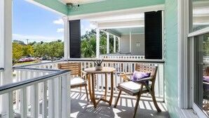 A covered balcony with a table and chairs, a potted plant, and a view of a street with parked cars and trees. - Crescent Cottage | Walk to Republic + Prohibition (Charleston)