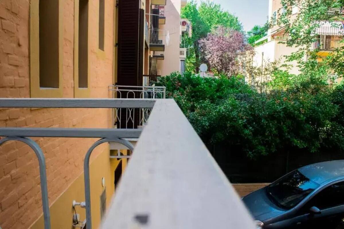 Un balcón estrecho con barandilla de metal, con vistas a una zona residencial que incluye un coche aparcado y un edificio con balcones.