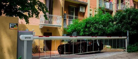 A gated entrance to a residential building with balconies, a car parked inside, and a small outdoor seating area.
