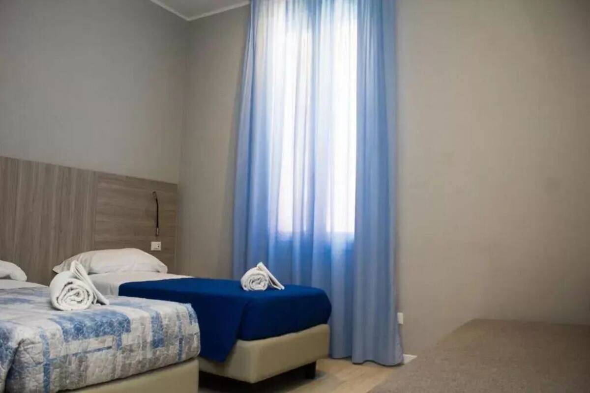 Habitación de hotel con dos camas, una con cubrecama azul y la otra con diseño estampado. Se ve un ventanal grande con cortinas azules, y un cabecero de madera al lado izquierdo de la habitación.