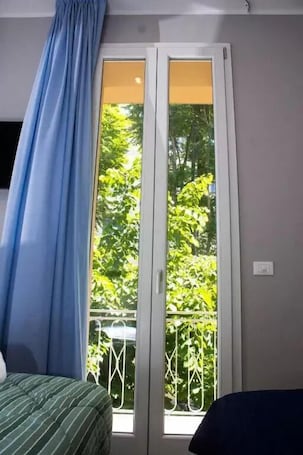 A room with a bed, a window with a view of greenery, and a curtain.