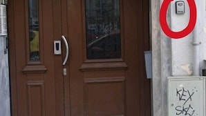 A brown wooden door with a glass panel and a small intercom. Graffiti is visible on the wall to the right. - Atelier Deluxe Apt Must (Thessaloniki)