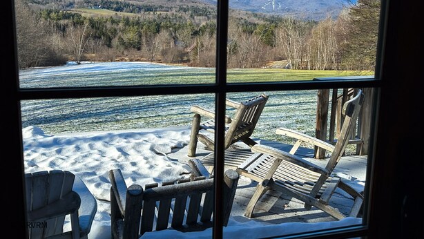 En vy genom ett fönster med en träterrass och Adirondack-stolar, med utsikt över ett snötäckt landskap och avlägsna berg.