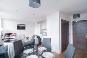 A modern living room with a dining area, grey sofas, a flat-screen TV, and a dark wooden door.