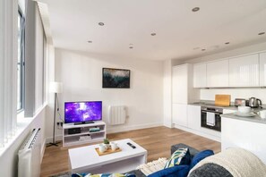 A modern living room with a flat-screen TV, a coffee table, a sofa, and a kitchen area with cabinets and appliances.
