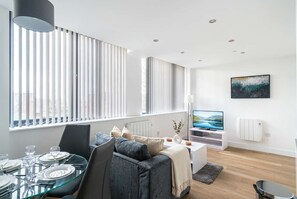A modern living room with a glass dining table, grey sofa, and a flat-screen TV.