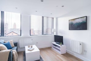A modern living room with a sofa, coffee table, TV, and large windows with blinds. - Comfortable 1BR Apartment in Manchester (Manchester)