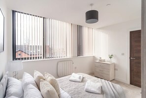 A modern bedroom with a large window, a bed with white bedding, a bedside table, and a dresser. - Comfortable 1BR Apartment in Manchester (Manchester)