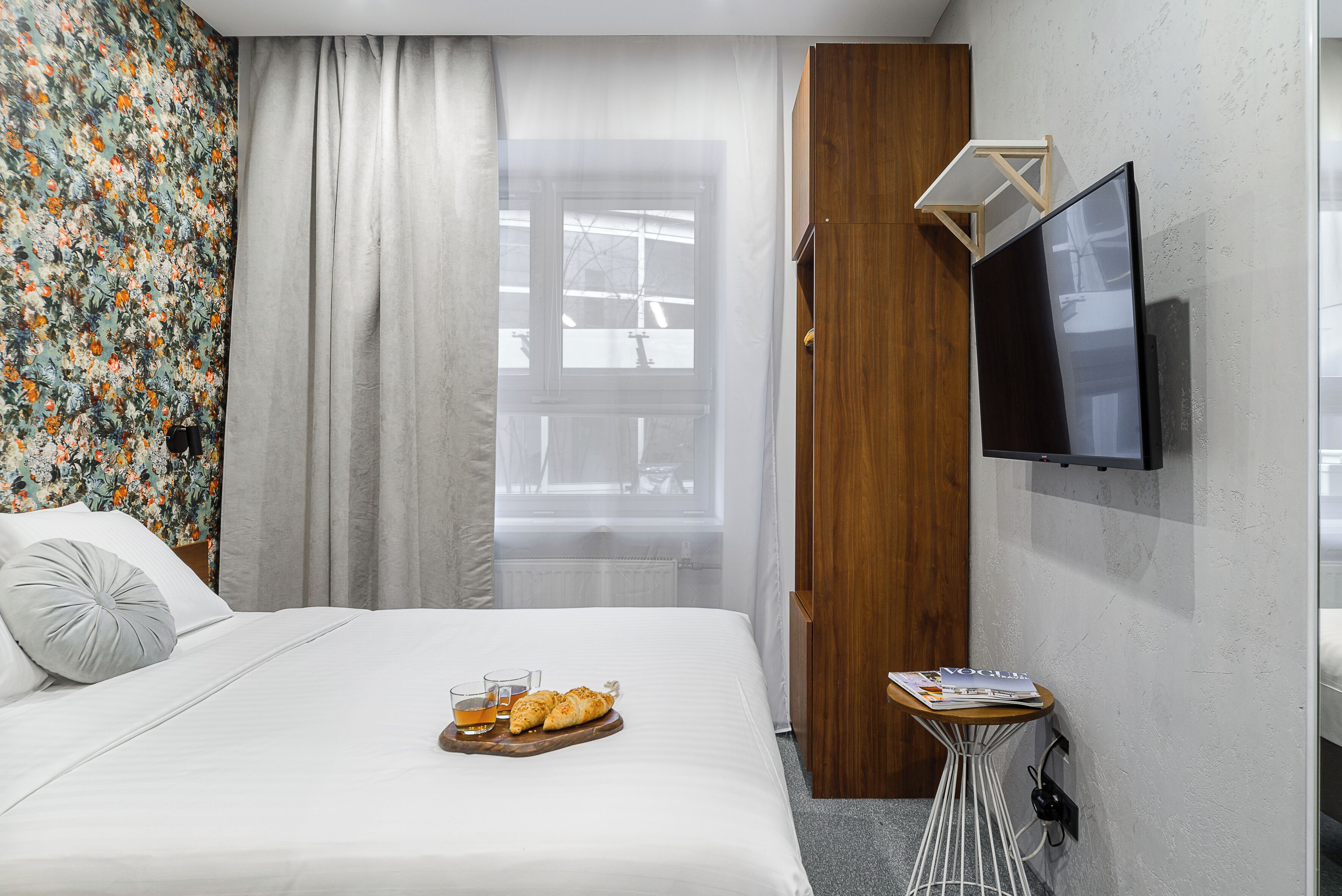 A hotel room with a bed, a TV mounted on the wall, a small table with a magazine, and a window with curtains.