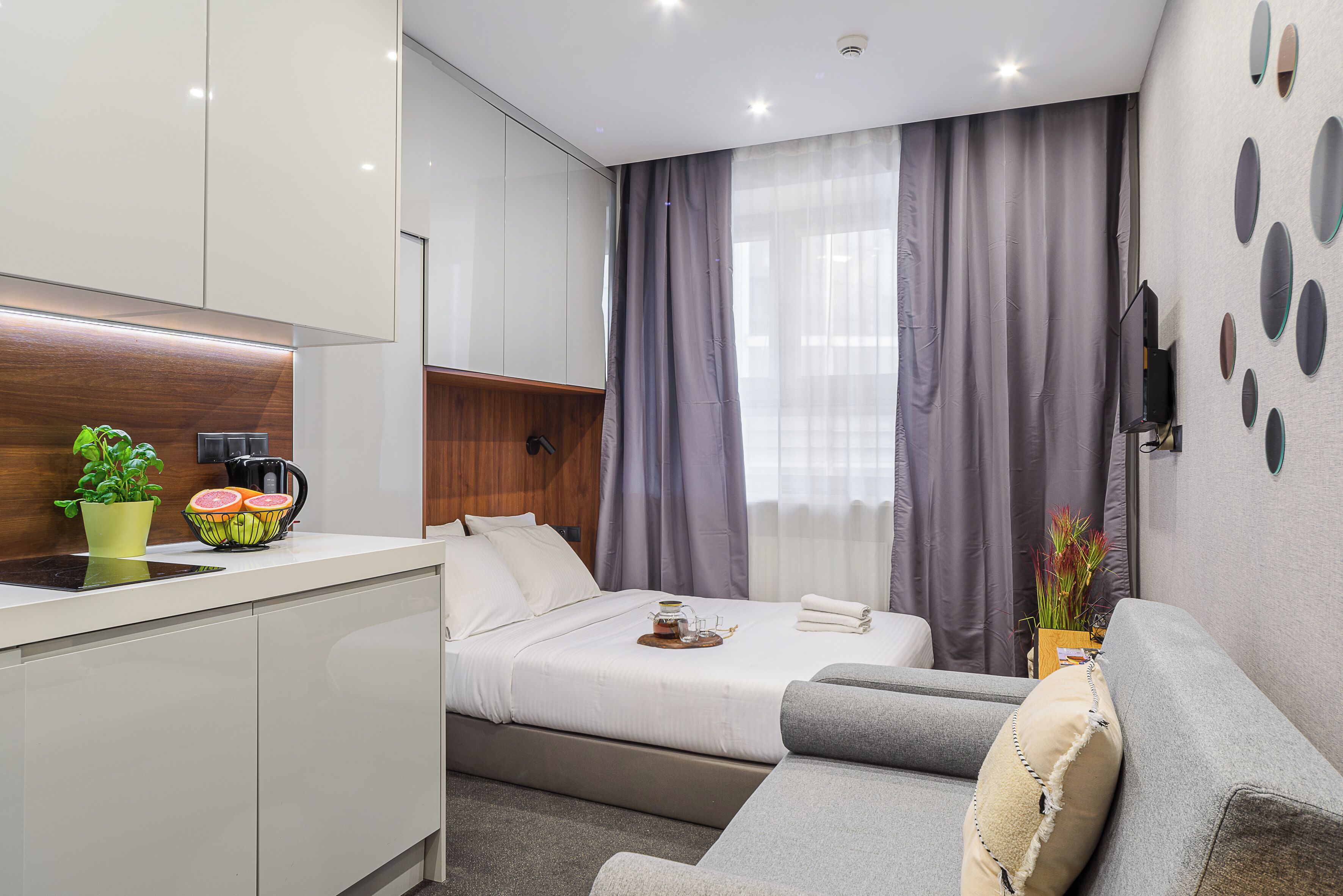 A modern hotel room with a kitchenette, a bed with a tray of breakfast, a sofa, and a wall with decorative mirrors.