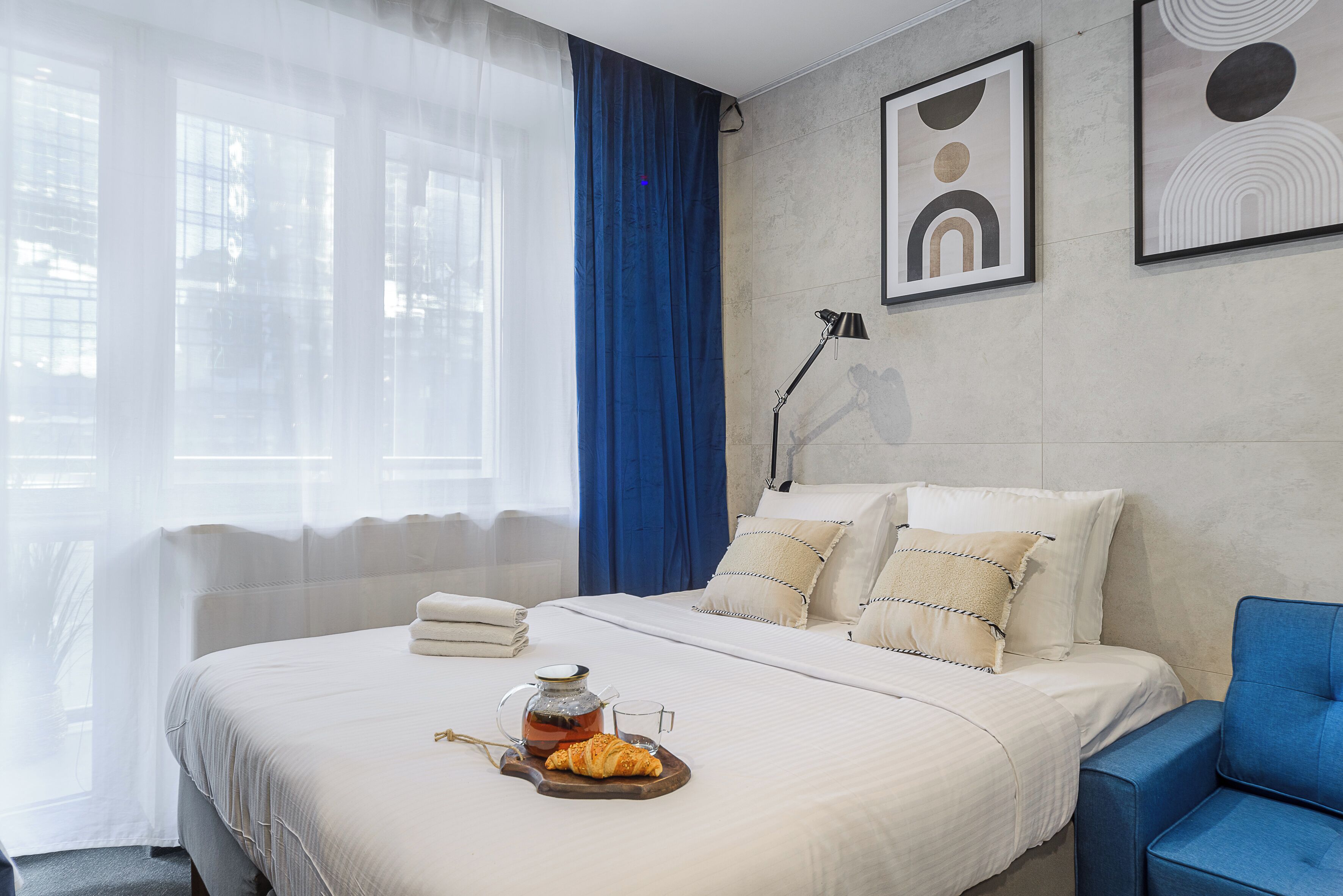 Un lit parfaitement fait, avec un plateau de petit-déjeuner, une lampe de chevet et une vue sur l’extérieur à travers de fines rideaux.
