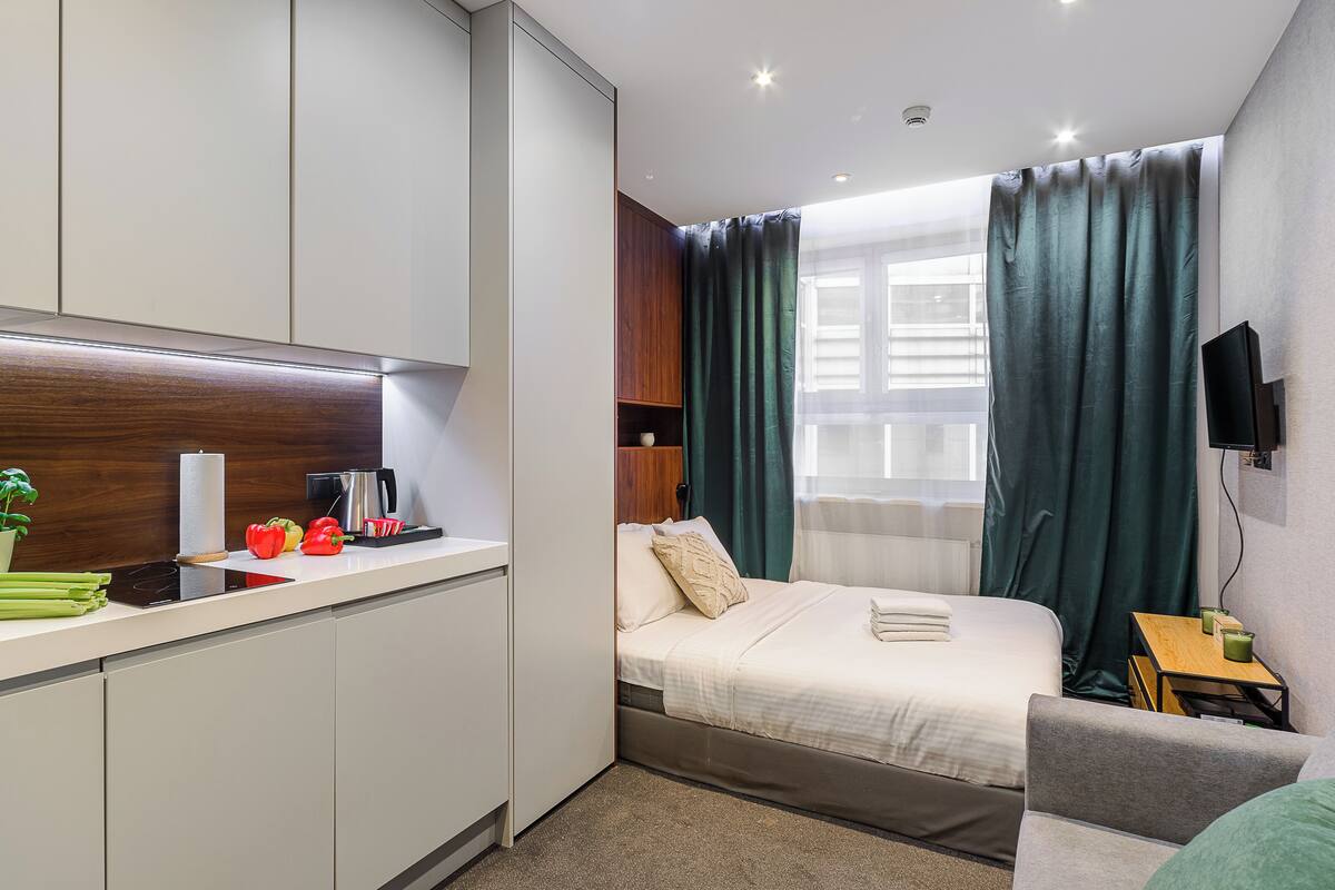 A compact hotel room with a kitchenette, a bed with a white comforter, a small table with a green cup, and a TV mounted on the wall.