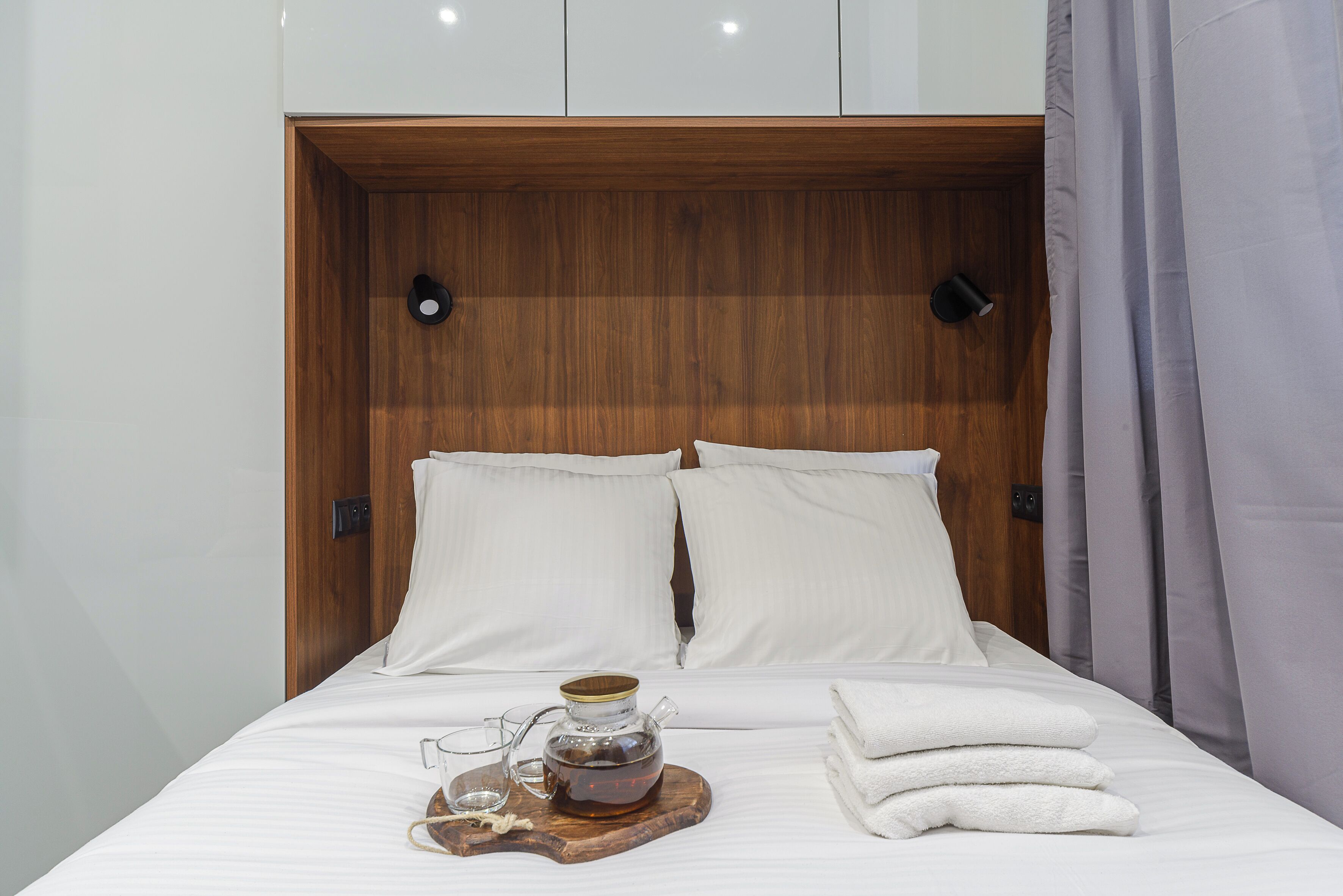 A neatly made bed with white linens, a glass teapot on a wooden tray, and folded white towels.